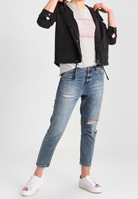 Svart kort jacka med huva, grå grafisk t-shirt, slitna blå denimjeans och vita sneakers med färgglada accenter.