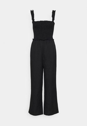 Mono sin mangas negro con cuerpo ajustado fruncido, tirantes con volantes y pantalones de pierna ancha sobre un fondo blanco liso.