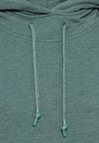 Primo piano di un tessuto di felpa verde che mostra il cappuccio e due cordini con punte di plastica annodate che pendono.
