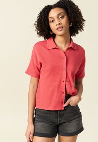 Kortärmad, korallfärgad skjorta med knappslå och strukturerat mönster. Har krage och träknappar, tillsammans med mörka jeansshorts.