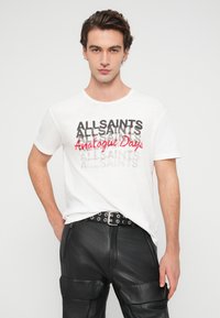 Άσπρο κοντομάνικο μπλουζάκι με στρωματοποιημένο κείμενο σε μαύρο και κόκκινο, που γράφει "ALLSAINTS Analogue Days." Συνδυάζεται με μαύρο δερμάτινο παντελόνι.