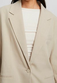 Beżowa marynarka z wyciętym kołnierzem, zapinana na jeden guzik, z kieszenią o subtelnej fakturze, noszona na jasnym topie.