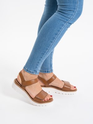 Pieds chaussés de sandales en cuir marron avec semelles blanches et vernis à ongles rouge, assortis à un jean skinny bleu sur fond blanc.