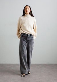 Cream, textured knit sweater with a high neck; gray wide-leg jeans; black and white printed heels; model stands against a plain background.
