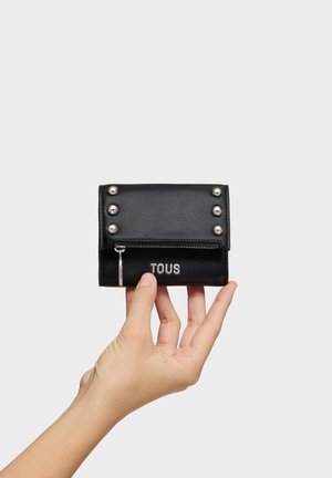 Hand holding a black leather wallet with metal studs, front zipper, and "TOUS" brand name on a plain white background.