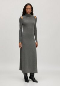 Femme portant une longue robe gris turtleneck avec des découpes aux épaules et des bottines noires à talons pointus, se tenant devant un fond uni.