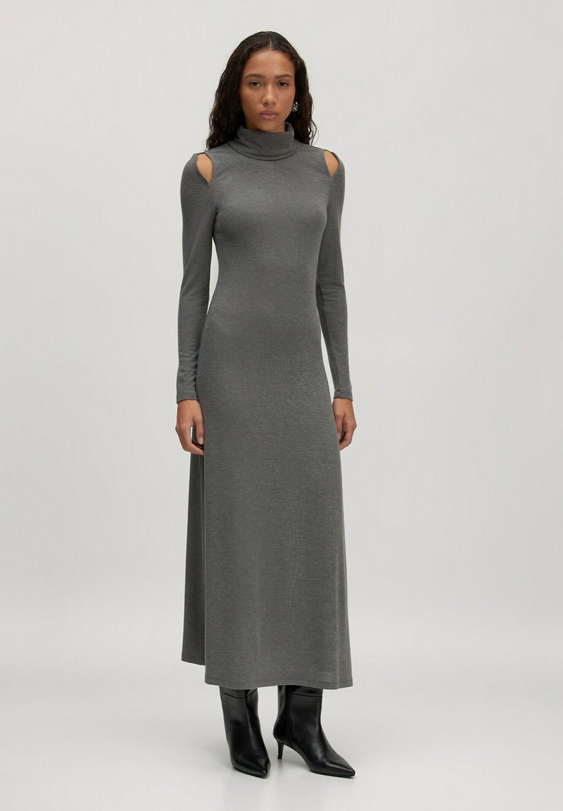 Femme portant une longue robe gris turtleneck avec des découpes aux épaules et des bottines noires à talons pointus, se tenant devant un fond uni.