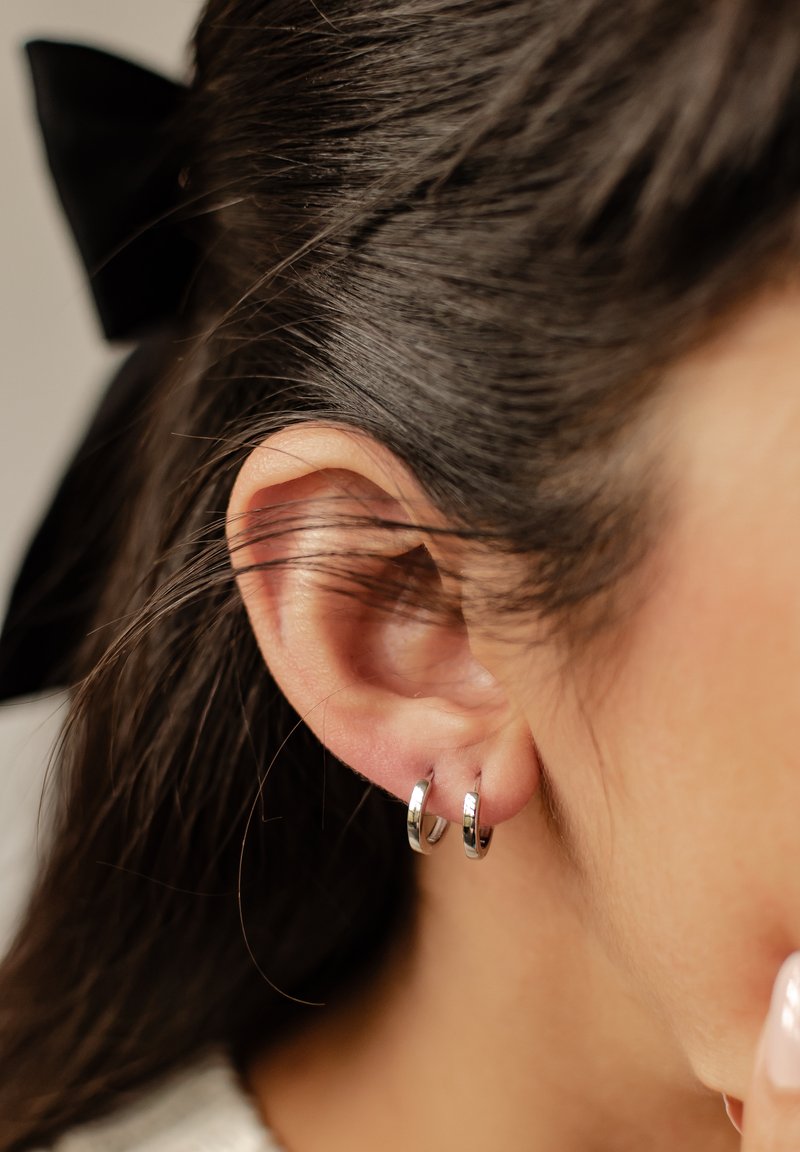 Pendientes de aro plateados reposando en una oreja, con una superficie brillante y suave y un diseño circular simple. El cabello oscuro y un lazo son visibles en el fondo.