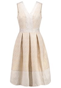 Robe cocktail beige sans manches avec col en V, jupe plissée et motif à pois brillants subtils sur tout le tissu.