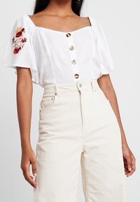 Blusa blanca de manga corta con escote cuadrado, que presenta grandes botones y un bordado floral colorido en el hombro izquierdo. Combinada con pantalones de pana beige.