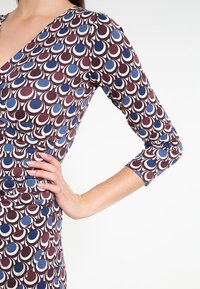 Långärmad klänning i ett mönstrat tyg med burgundy, blå och vita cirklar; figursydd design med v-ringning och ruching på sidan.