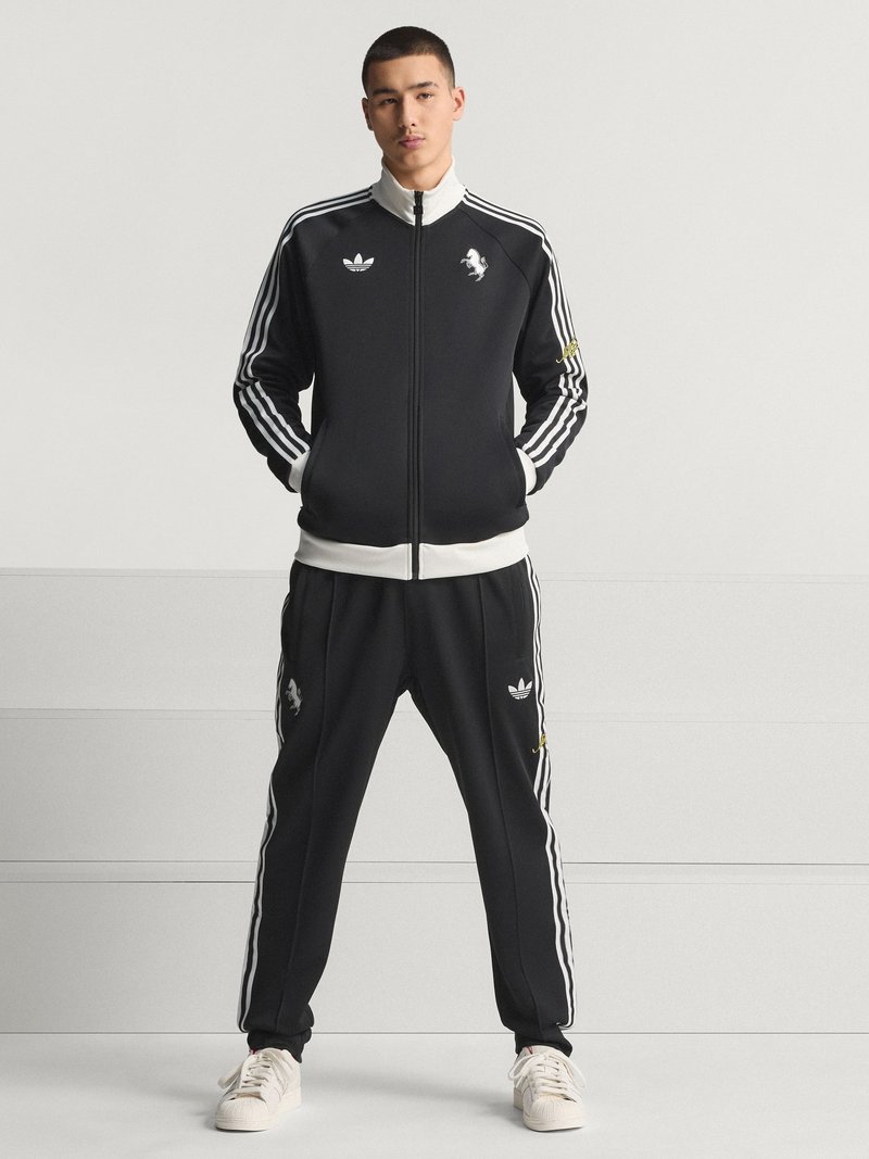 Survêtement noir avec un design à trois bandes blanches et un logo de lion. La veste possède un col montant et une fermeture éclair, tandis que le pantalon est doté de poches latérales et de poignets.