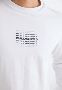 Tricou alb cu mânecă lungă, prezentând un print grafic cu textul "KARL LAGERFELD" în negru, aranjat în straturi suprapuse, transparente.