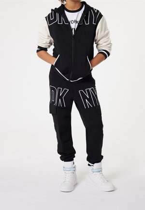 Černá mikina na zip s bílými prvky a velkými písmeny "NY", v kombinaci s černými cargo kalhotami s bílým nápisem a bílými sneakers s vysokým střihém.