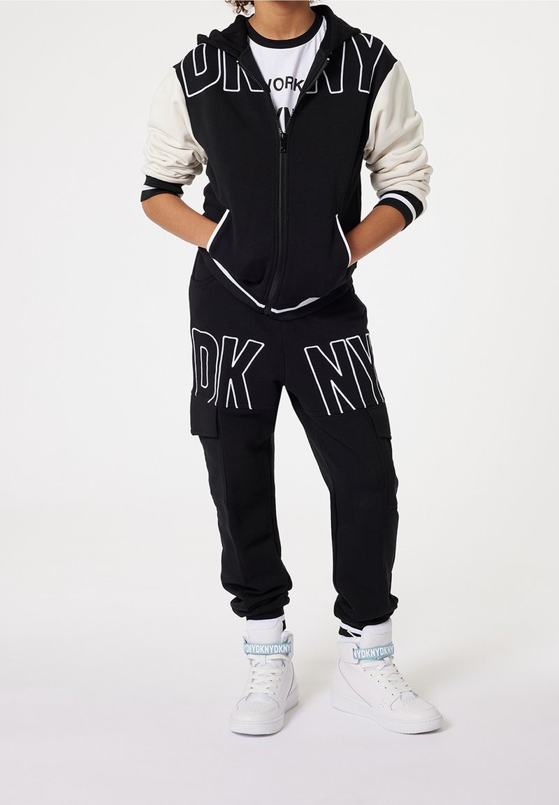 Černá mikina na zip s bílými prvky a velkými písmeny "NY", v kombinaci s černými cargo kalhotami s bílým nápisem a bílými sneakers s vysokým střihém.