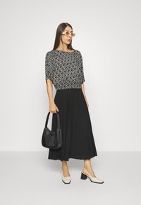 Saia plissada preta com uma blusa cinza estampada. Modelo segura uma bolsa preta, usa sapatos de salto quadrado bege e óculos de sol brancos oversized.