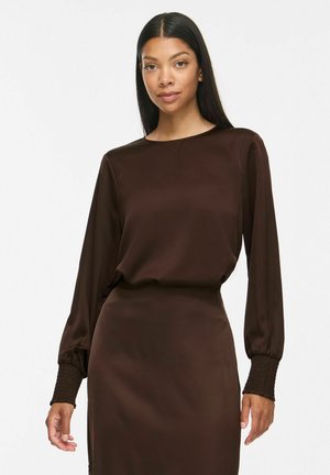 Femme aux longs cheveux noirs portant une robe marron à manches longues avec des poignets froncés, debout devant un fond blanc uni.