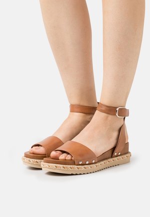 Sandales en cuir marron avec une large lanière au niveau des orteils et une lanière de cheville réglable, ornées d'un accent en jute tissé et de petits clous métalliques.