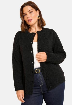 Woman wearing a black textured button-up cardigan over a white top and dark blue jeans with a black belt featuring a gold circular buckle.