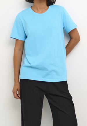 Persona con una camiseta de manga corta azul claro lisa y pantalones negros holgados, de pie sobre un fondo blanco.