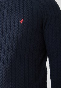 Pull en maille bleu marine avec un motif en relief, arborant un petit logo de rouge-gorge sur la poitrine, fabriqué en tissu doux et texturé.