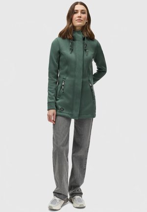 Groene fleecejas met een hoge kraag, ritszakken en toggle-accenten. Gecombineerd met grijze broek en lichtgekleurde schoenen.