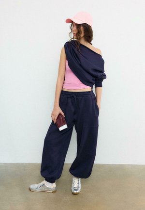 Vrouw met een roze pet, roze tanktop met een donkerblauwe trui losjes over één schouder gedragen, donkerblauwe joggingbroek, zilveren sneakers, houdend een portemonnee met kaartjes.