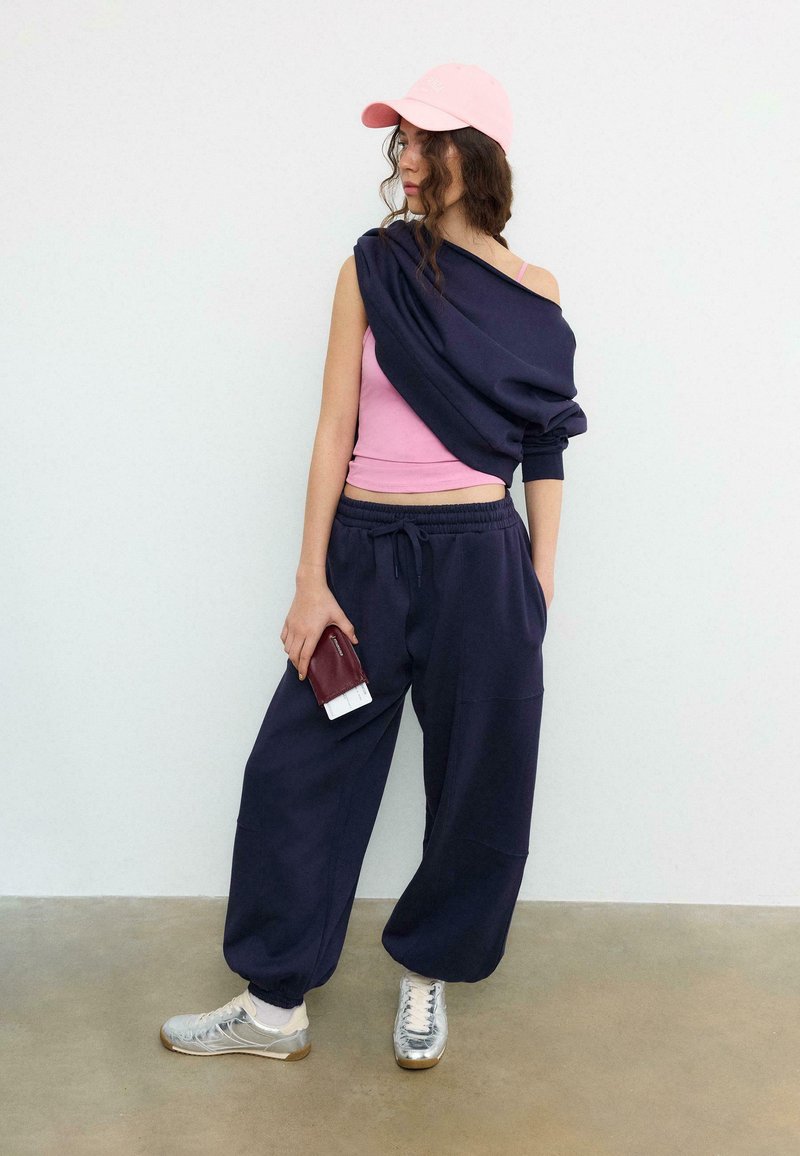 Žena s ružovou čiapkou, ružovým tielkom a tmavomodrou mikinou prehodenou cez jedno rameno, tmavomodré tepláky, strieborné tenisky, drží peňaženku s lístkami.