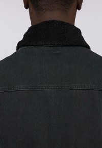 Giubbotto in denim scuro con colletto in shearling nero e morbido, con cuciture visibili e un'etichetta sul retro vicino al collo.