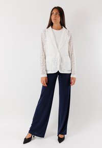 Blazer bianco in pizzo con maniche lunghe, abbinato a pantaloni a gamba larga blu navy. Il blazer presenta due tasche frontali e una chiusura a un bottone.