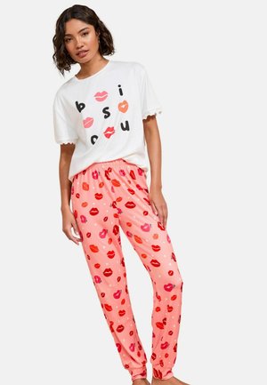 Žena v bielom tričku s dizajnom pier a písmen, v kombinácii s ružovými pyžamovými nohavicami s potlačou červených a ružových pier.