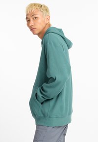 Sweat à capuche bleu sarcelle avec texture côtelée, poche avant et poignets ajustés. Modèle présenté de profil, aux cheveux blonds courts et fond neutre.