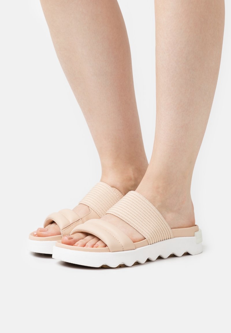 Pieds portant des sandales beige à enfiler avec de larges brides en tissu et des semelles blanches épaisses à motif strié sur fond blanc.
