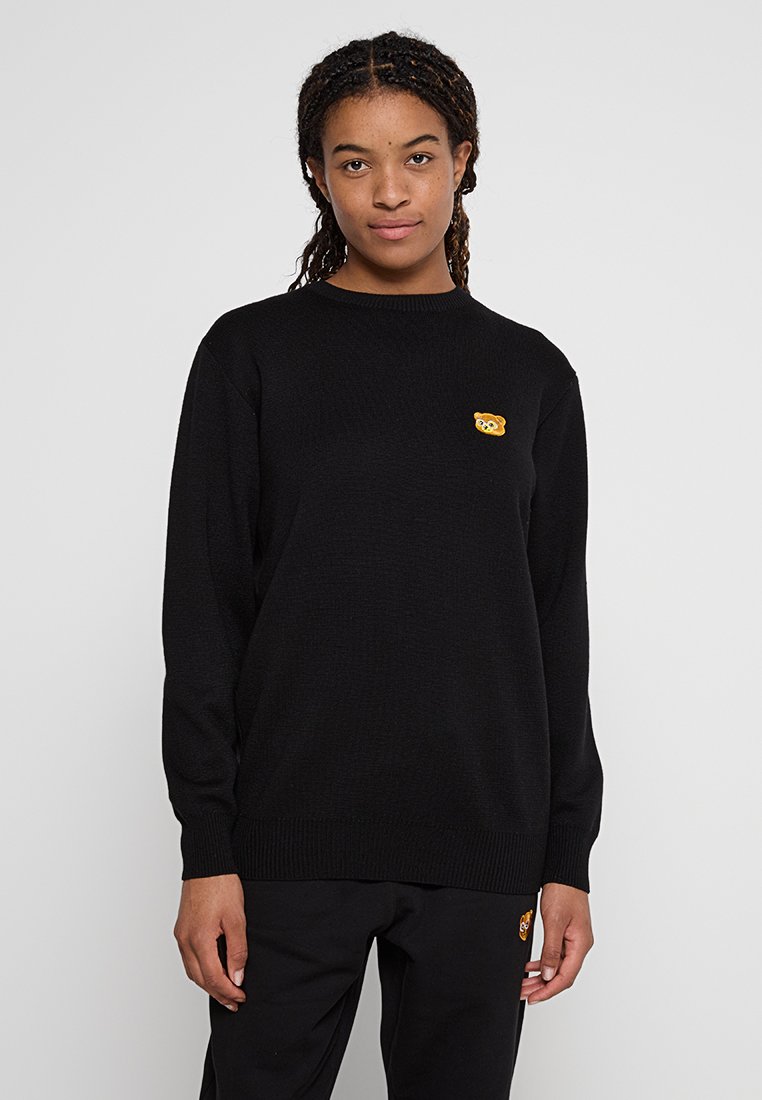 Pull noir à col rond, à manches longues, avec un petit patch d'ours jaune sur la poitrine. Texture en tricot lisse, design décontracté.