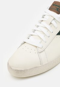 Hvit lær sneakers med perforert tå, grønn aksentstripe og brun logo-patch. Har flate hvite snøringer og teksturert gummisåle.