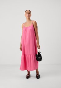 Ροζ αμάνικο φόρεμα με πτυχώσεις στον λαιμό, tiered hem και υφή υφάσματος. Το μοντέλο κρατάει μια μικρή μαύρη τσάντα με λαβή από πέρλες.