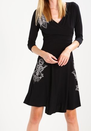 Frau trägt ein schwarzes knielanges Kleid mit weißen Blumenmustern, dreiviertel Ärmeln und V-Ausschnitt, steht vor einem weißen Hintergrund.