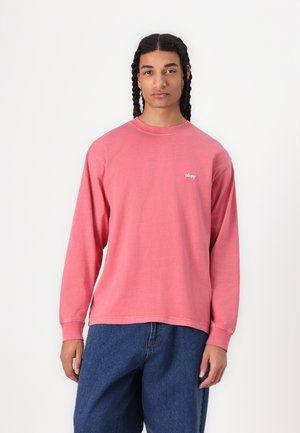 Light pink long-sleeve shirt made from soft material, with a crew neck and a small white "obey" logo on the chest, paired with loose blue jeans.