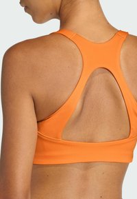 Sujeción deportiva naranja con diseño de espalda en forma de racerback, dotada de un corte en forma de llave y una textura suave. Detalles de costura a lo largo de los bordes.
