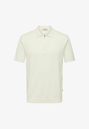 Fiatal férfi, fekete dzsekit, krémszínű félcipzáras pulóvert, szürke nadrágot és fekete cipőt visel, egy kézzel a zsebében áll.