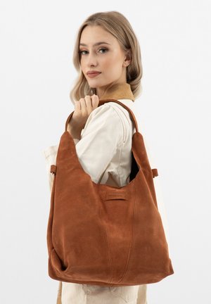 Donna bionda con giacca chiara che tiene una grande borsa a tracolla in pelle scamosciata marrone con patch logo su sfondo bianco.