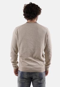 Uomo con capelli ricci che indossa un maglione beige e jeans blu con il marchio "DS2", in piedi di spalle su uno sfondo bianco.