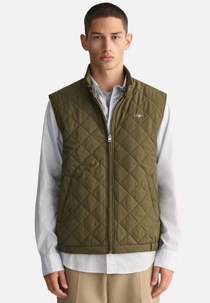 Gilet matelassé vert olive avec un col haut, une fermeture éclair à l'avant et un logo brodé sur la poitrine, superposé à une chemise striped bleu clair.
