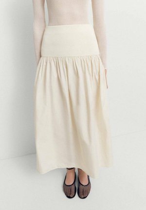 Femme portant une longue jupe crème avec taille froncée, un haut à manches longues beige côtelé et des ballerines noires en résille, debout sur un sol blanc.