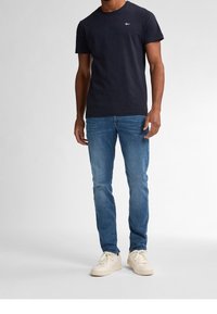 Man som har på sig en mörk marinblå t-shirt med kort ärm, blå slim-fit jeans och off-white sneakers, står framför en enfärgad ljus bakgrund.