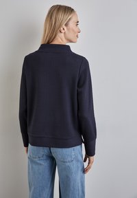 Granatowy, prążkowany sweter z wysokim kołnierzem i długimi rękawami; ma lekko skrócony krój oraz teksturowane wykończenie. Noszony z jasnoniebieskimi dżinsami.