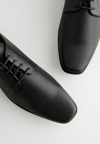 Zwarte synthetische dresschoenen met een gestructureerd oppervlak, voorzien van subtiele rode stiksels, ovale veters en een puntige neus.