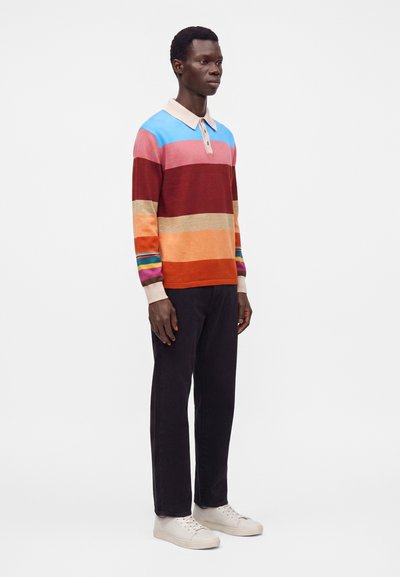 Homme debout portant un polo à manches longues rayé multicolore, un pantalon noir et des baskets blanches sur fond blanc.