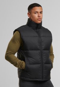 Schwarze Pufferweste mit hohem Kragen, Frontreißverschluss und zwei seitlichen Taschen. Glatte Textur mit gesteppten horizontalen Feldern.