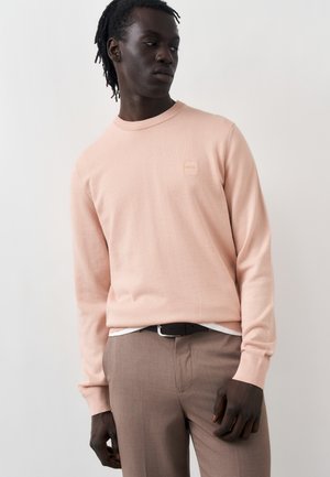 Uomo con pelle scura e capelli intrecciati che indossa un maglione rosa chiaro e pantaloni beige, in piedi davanti a uno sfondo chiaro e uniforme.